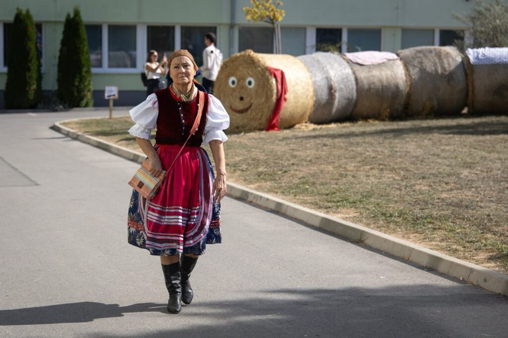 Žena v kroji pred ozdobenými balíkmi slamy v areáli školy